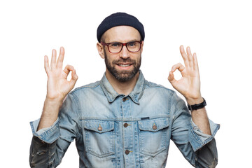 Naklejka premium Cheerful bearded male in fashionable clothing, makes ok sign, being satisfied with something, isolated over pink studio background. Unshaven middle aged man gestures indoor, shows his approval