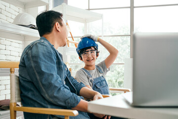 Cheerful Asian carpentry father discusses and teaches his son who wears a safety helmet about...