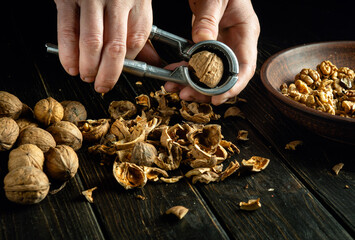 Peeling walnuts with a nutcracker in hands of a cook. Peeled walnuts in a plate and on the black kitchen table