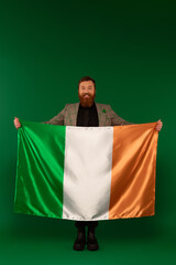 Full length of positive bearded man holding Irish flag on green background.