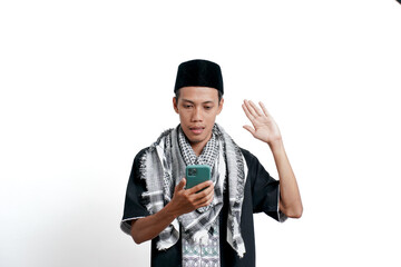 A religious Muslim Asian man wearing a turban, Muslim clothes and a cap, holding a smartphone like he's on a video call, and is shocked. Isolated on white background.