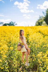 Obraz na płótnie Canvas girl is standing in a yellow field of rapeseed. A woman with a bouquet of flowers in a bag and a hat