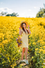 girl in a hat walks in a yellow field of rapeseed. A woman with a bouquet of flowers in her bag