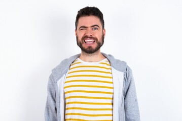 Coquettish caucasian man wearing casual sportswear over white wall smiling happily, blinking at...