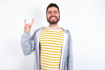 Naklejka premium caucasian man wearing casual sportswear over white wall doing a rock gesture and smiling to the camera. Ready to go to her favorite band concert.