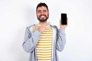 Smiling caucasian man wearing casual sportswear over white wall Mock up copy space. Pointing index finger on mobile phone with blank empty screen