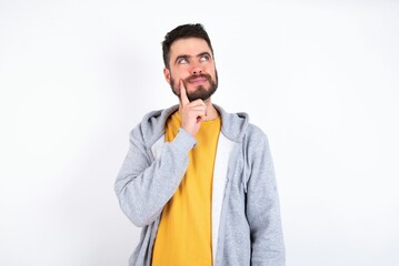 Thoughtful caucasian man wearing casual sportswear over white wall holds chin and looks away pensively makes up great plan