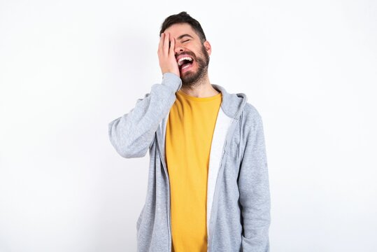 caucasian man wearing casual sportswear over white wall makes face palm and smiles broadly, giggles positively hears funny joke poses
