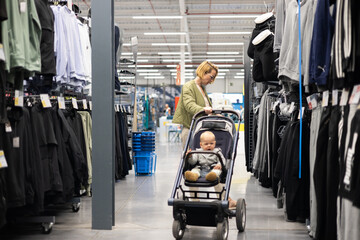 Casualy dressed mother choosing sporty shoes and clothes products in sports department of supermarket store with her infant baby boy child in stroller - obrazy, fototapety, plakaty