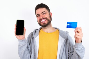 caucasian man wearing casual sportswear over white wall opened bank account, holding smartphone and credit card, smiling, recommend use online shopping application