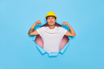 Self confident asian male worker raises arms shows muscles and power to work gestures thumbs up wears protective hardhatposes through blue torn paper hole
