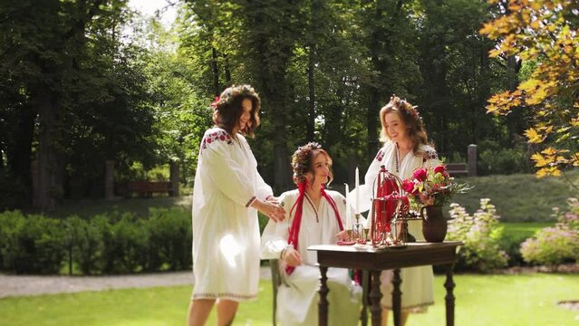 Three women stand next to beauty table and admire each other . Girls wear Ancient ethno Ukrainian embroidered shirt dress and wreath of flowers. Close up Medium shot