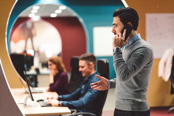 Side view of a man talking on the phone, while at the office of a start up company, his colleagues working by their desks in the background
