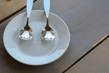 One clean white plate is standing on the wooden table with two metal spoons on it. Two metal spoons on the clean white plate. Ready to eat something