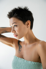 Fototapeta na wymiar young tattooed woman in strapless top holding hand near head and looking away isolated on grey.