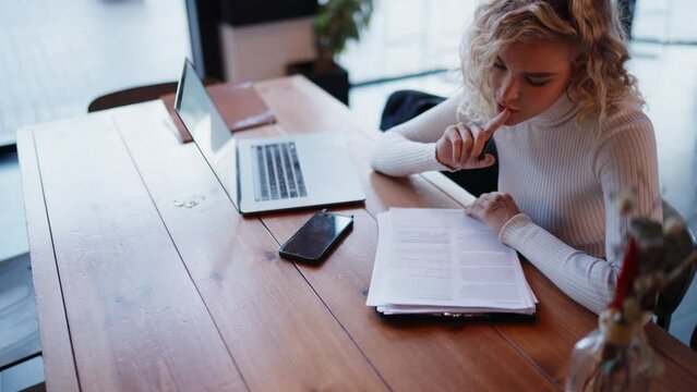 Caucasian young woman making notes while working in coffee shop with laptop computer. Blonde hair female fills Model Release for stock photography while sitting at wooden table with netbook and phone