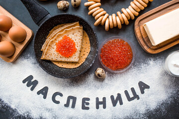 Russian traditions. Russian holiday Maslenitsa. A stack of pancakes, red caviar, bagels, bagels, a wooden spoon, eggs and butter. The Russian phrase Maslenitsa is written with flour. View from above.