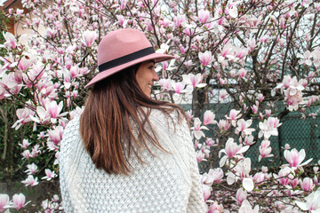 Girl with a pink hat posing next to flowers in bloom. Beautiful magnolia tree in spring. Girl profile smiling with long hair and white jumper. © Elsa
