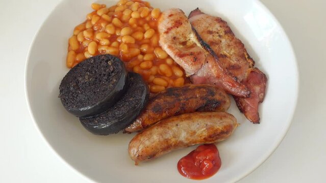Tomato ketchup being added to a traditional English breakfast of cumberland sausages, back bacon black pudding and baked beans, but no egg, on a white plate.  Real time, zoom out