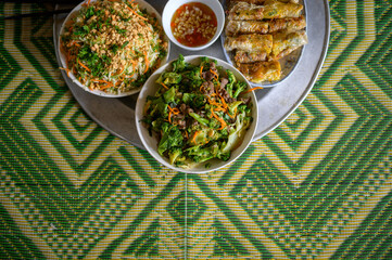Top view of appetizing Nem ran served on ceramic plate near green salads with chickpeas and broccoli and small bowl with nuoc cham on green ornamental tablecloth