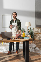 Smiling carpenter holding smartphone near tools and devices in workshop.