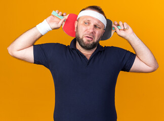 scared adult slavic sporty man wearing headband and wristbands holding table tennis rackets behind...