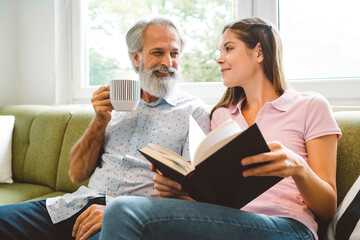 Smiling senior man enjoys spending his time together with his granddaughter, young woman sitting next to him holding a book 