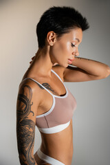 Fototapeta premium side view of brunette woman with tattooed body posing in bra with hand near neck on grey background.