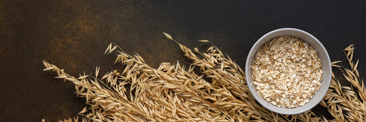 Oat flakes in a bowl and ears of oats on a brown background, top view