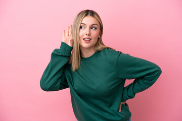 Young caucasian woman isolated on pink background listening to something by putting hand on the ear