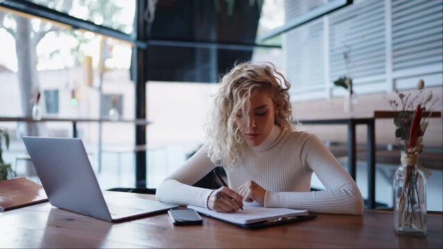 Caucasian young woman making notes while working in coffee shop with laptop computer. Blonde hair female model fills Model Release for stock photography while sitting at wooden table with netbook