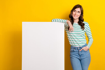 Fototapeta na wymiar Photo of cheerful woman curly hairstyle striped shirt stand near white billboard look empty space isolated on yellow color background