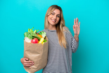 Young Uruguayan woman holding a grocery shopping bag isolated on blue background saluting with hand...