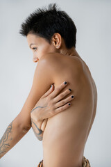 Fototapeta na wymiar side view of shirtless tattooed woman covering breast with hand isolated on grey.
