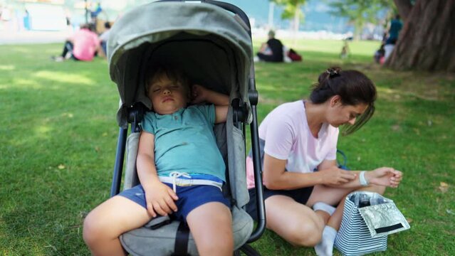 Child asleep inside stroller at park with mother sitting at grass. Family weekend lifestyle moment. Little boy napping