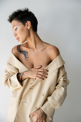 Fototapeta na wymiar sensual tattooed woman with bare shoulders covering breast with hand while posing in oversize blazer isolated on grey.