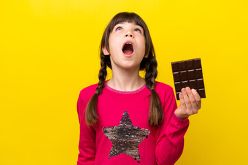 Little caucasian girl with chocolat isolated on yellow background looking up and with surprised expression