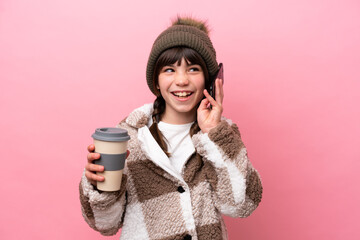 Little caucasian girl with winter jacket isolated on pink background holding coffee to take away and a mobile