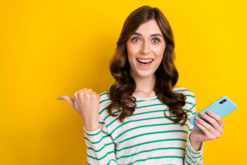 Photo portrait of lovely young lady hold gadget excited point empty space wear trendy striped outfit isolated on yellow color background
