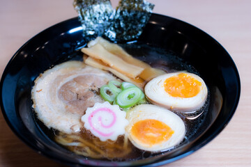 Soy sauce ramen in a black cup topped with chashu pork, Naruto fish balls, soy-sauce pickled eggs, menma bamboo shoots and seaweed. Ramen is a distinctive Japanese style noodle soup.
