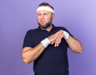 unpleased adult slavic sporty man wearing headband and wristbands holding hands together and looking at side isolated on purple background with copy space