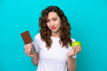 Young caucasian woman isolated on blue background taking a chocolate tablet in one hand and an...