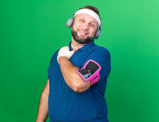 smiling adult slavic sporty man on headphones wearing headband wristbands and phone armband puts...