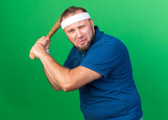 scared adult slavic sporty man wearing headband and wristbands holding bat isolated on green background with copy space