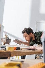 Brunette carpenter holding sandpaper and blowing on wooden board in workshop.