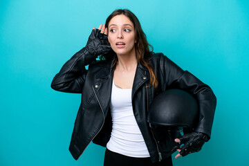 Young caucasian woman with a motorcycle helmet isolated on blue background listening to something by putting hand on the ear