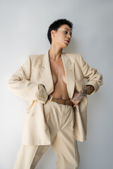 tattooed woman wearing elegant oversize suit on sexy and shirtless body while standing with hands on waist on grey background.