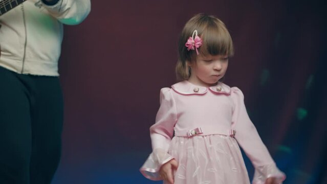 Little girl on stage in pink vintage dress, she dances, her father plays acoustic guitar. Color music is shining. Performance on stage. Early development of children