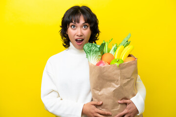 Young Argentinian woman holding a grocery shopping bag isolated on yellow background looking up and with surprised expression