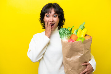 Young Argentinian woman holding a grocery shopping bag isolated on yellow background with surprise and shocked facial expression